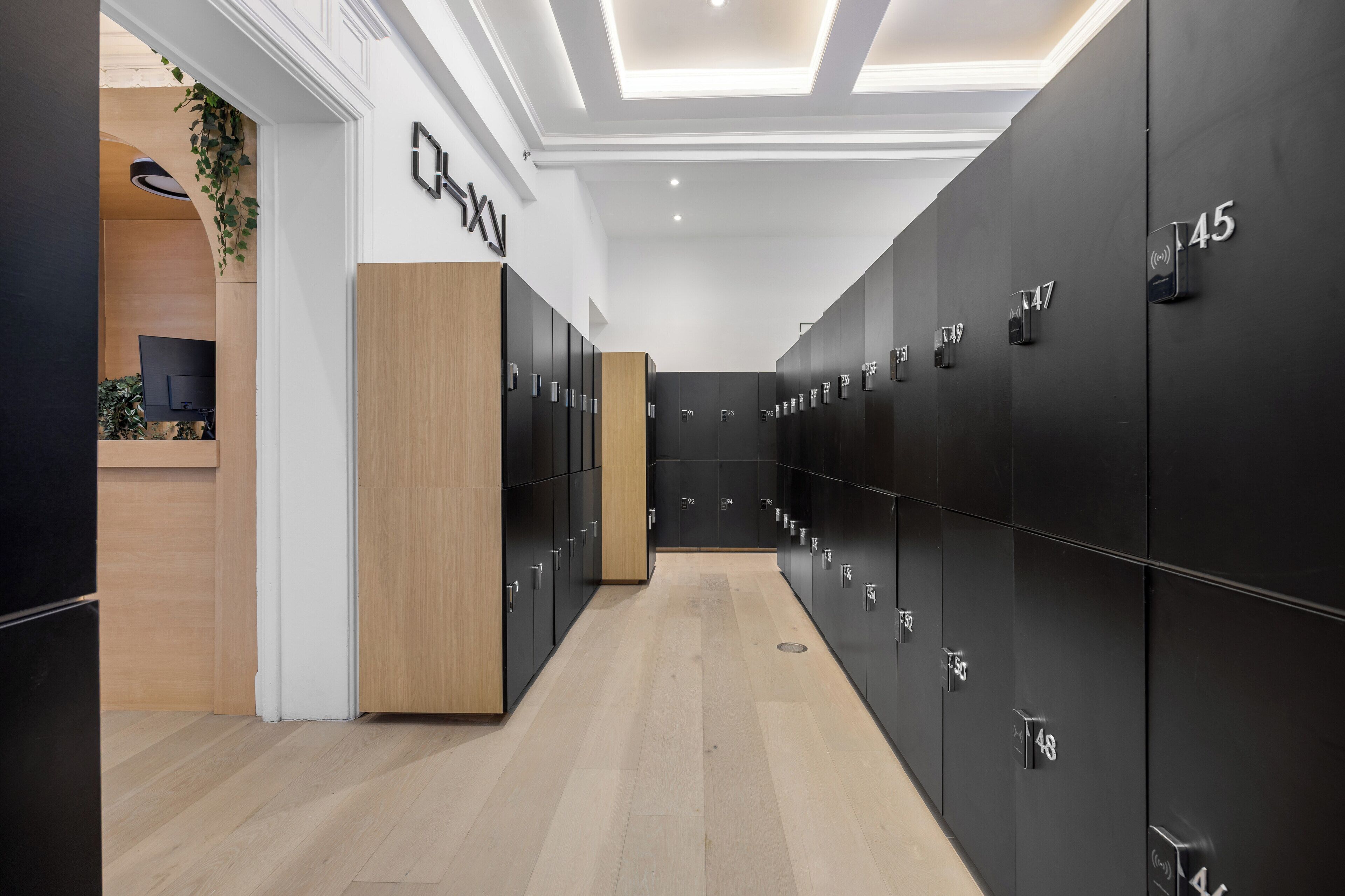A modern hotel lobby with a wooden reception desk, black lockers numbered 45-49, and a ceiling with recessed lighting.