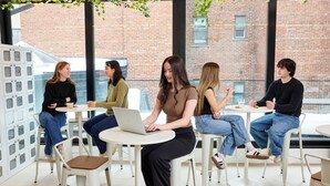 Un grupo de personas sentadas en mesas redondas con computadoras portátiles, en una habitación con ventanas amplias y una pared de ladrillo al exterior.
