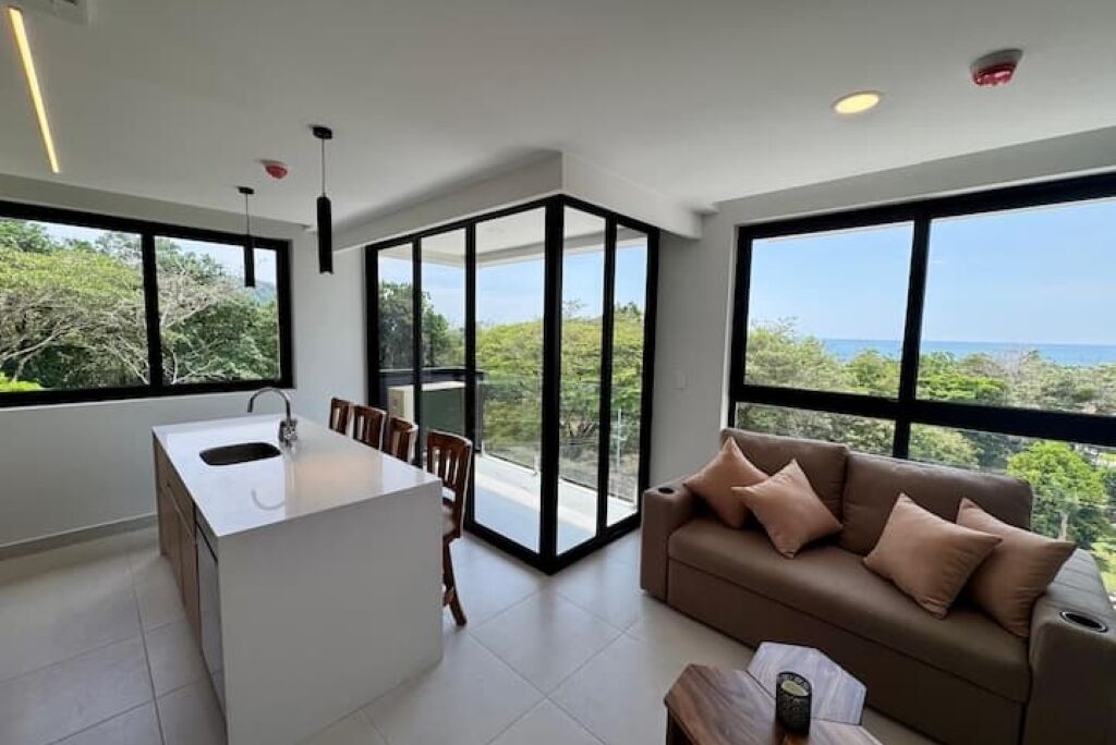 Una cocina moderna con una isla central, un comedor con sillas, un sofá con cojines y ventanas amplias que ofrecen vistas al exterior.