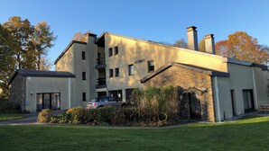 A modern multi-story building with a stone facade, a car parked in front, and a well-maintained lawn. - Quiet gîte between hills and forest. Sauna and pets welcome. (Bièvre)