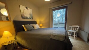 A bedroom with a large bed, a nightstand with a lamp, a chair, a painting on the wall, and a window with a view of trees. - Quiet gîte between hills and forest. Sauna and pets welcome. (Bièvre)