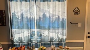 A dining area with a wooden table, two cushioned benches, a window with a forest-themed curtain, and a small table with snacks and a box labeled "SMORES & STATION".