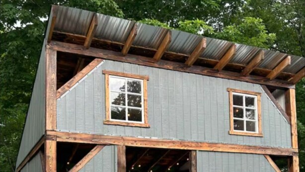 A small, rustic cabin with a gabled roof and wooden beams, featuring two windows and a door leading to an interior with a bed and various decorations.