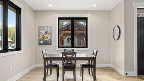 A dining area with a wooden table and chairs, a painting on the wall, a window with a view of a building, and a clock on the wall. - Newly Built 1BR Cottage - Rock Creek Cottages #5 (Red Lodge)