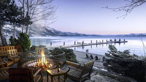 Ein Sitzbereich im Freien am Seeufer mit Feuerstelle, Holzmöbeln und Blick auf einen Steg, der in einen ruhigen See führt, mit schneebedeckten Bergen im Hintergrund.