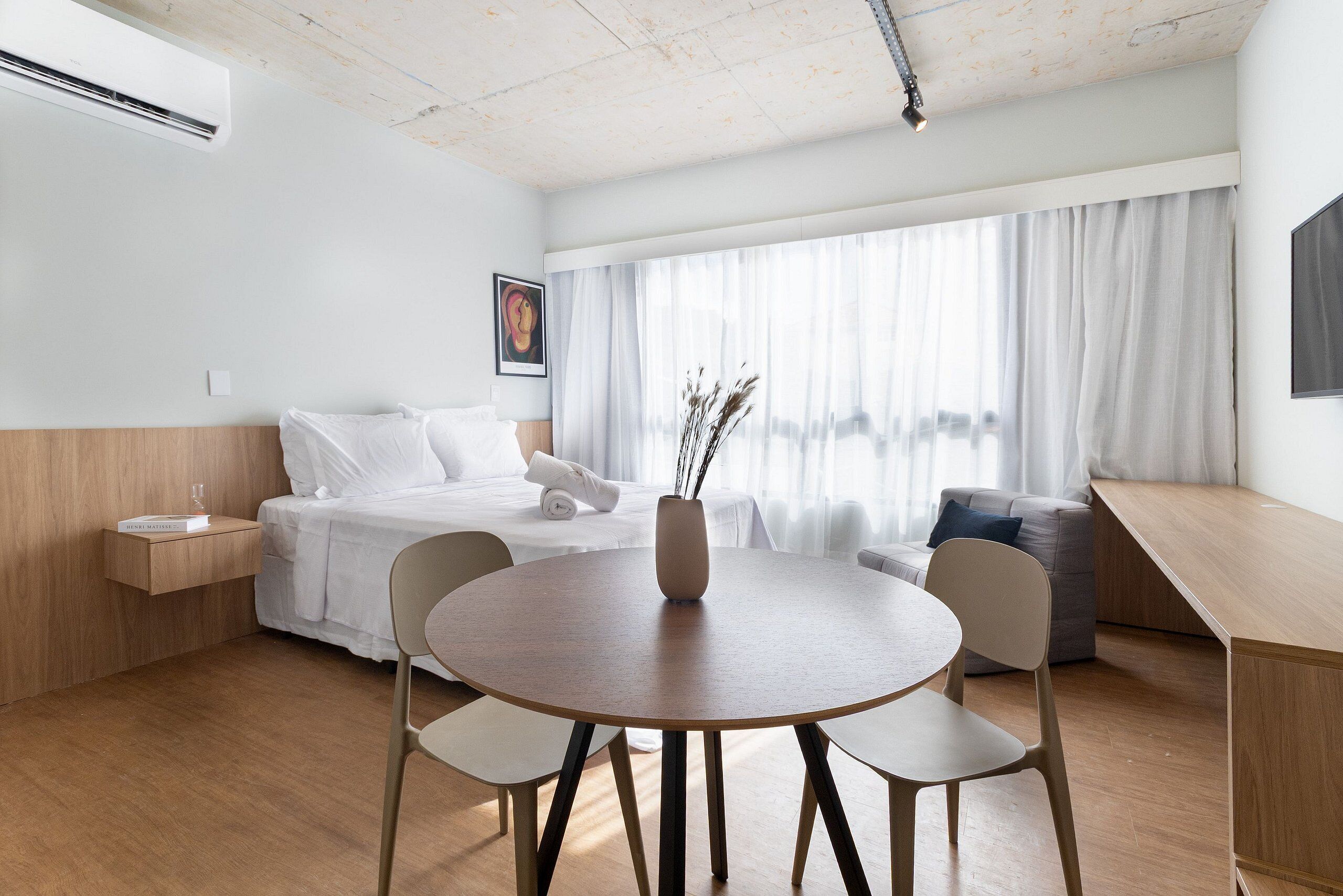 Una habitació d'hotel moderna amb terra de fusta, una taula rodona amb cadires, un llit amb roba de llit blanca, una taula de nit amb un got, i una finestra gran amb cortines transparents.