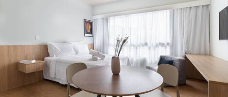 Una habitació d'hotel moderna amb terra de fusta, una taula rodona amb cadires, un llit amb roba de llit blanca, una taula de nit amb un got, i una finestra gran amb cortines transparents.
