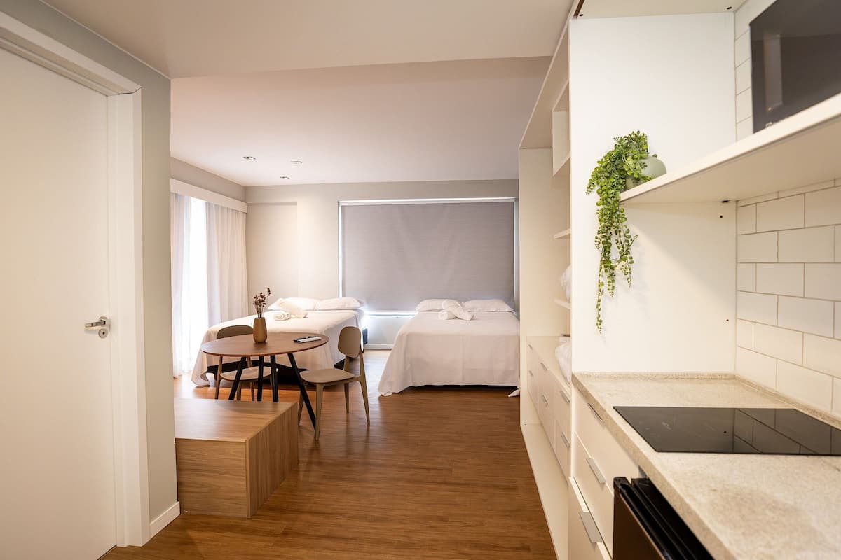 A modern, compact hotel room with a wooden floor, a dining area with a table and chairs, a bed with white bedding, and a kitchen area with a countertop and a hanging plant.