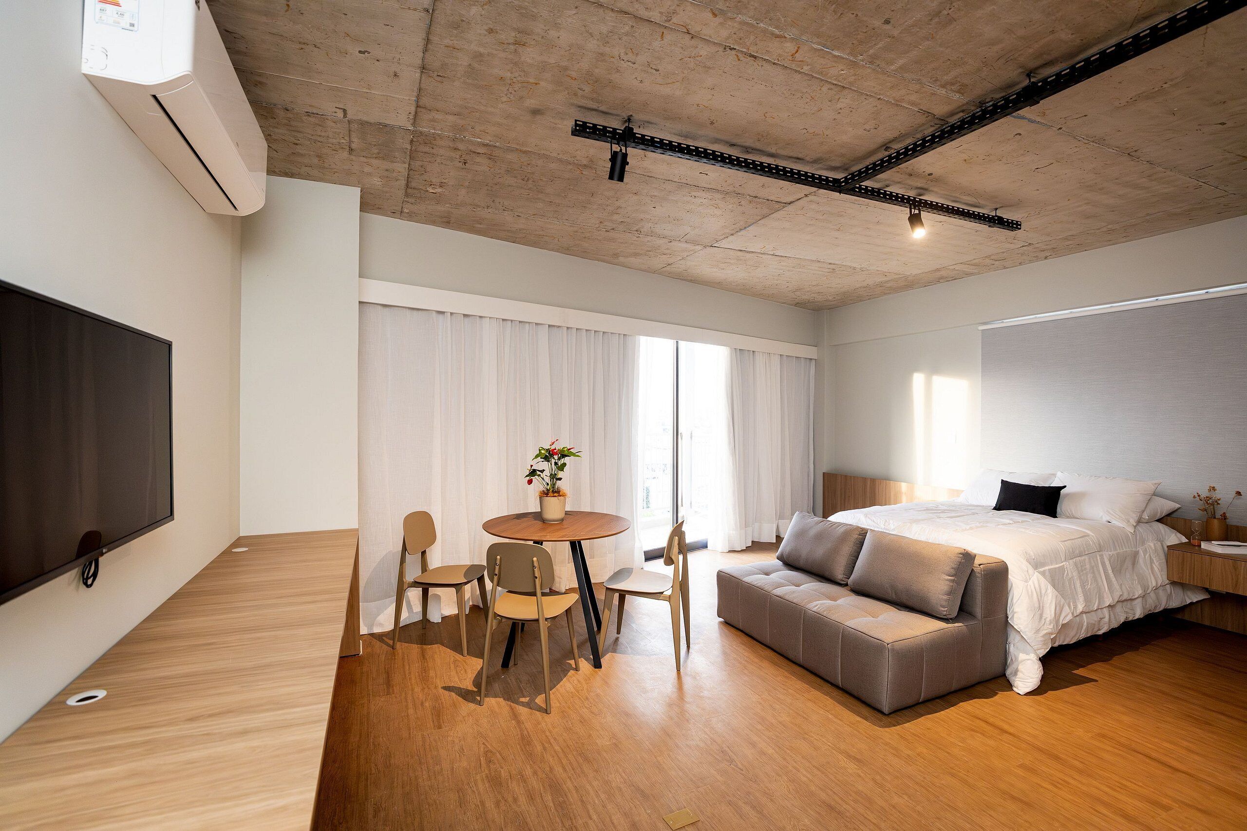 A modern hotel room with a wooden floor, a bed with a grey upholstered headboard, a round dining table with chairs, a flat-screen TV mounted on the wall, and a ceiling with exposed beams and lighting fixtures.