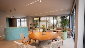 Un espace repas moderne avec une table ronde en bois entourée de chaises en osier, un comptoir de cuisine avec des carreaux bleus et de grandes fenêtres offrant une vue sur l’extérieur.
