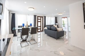 Ein modernes Wohnzimmer mit einem grauen Sofa, einem Glastisch mit Stühlen und einem großen Fenster mit Vorhängen.