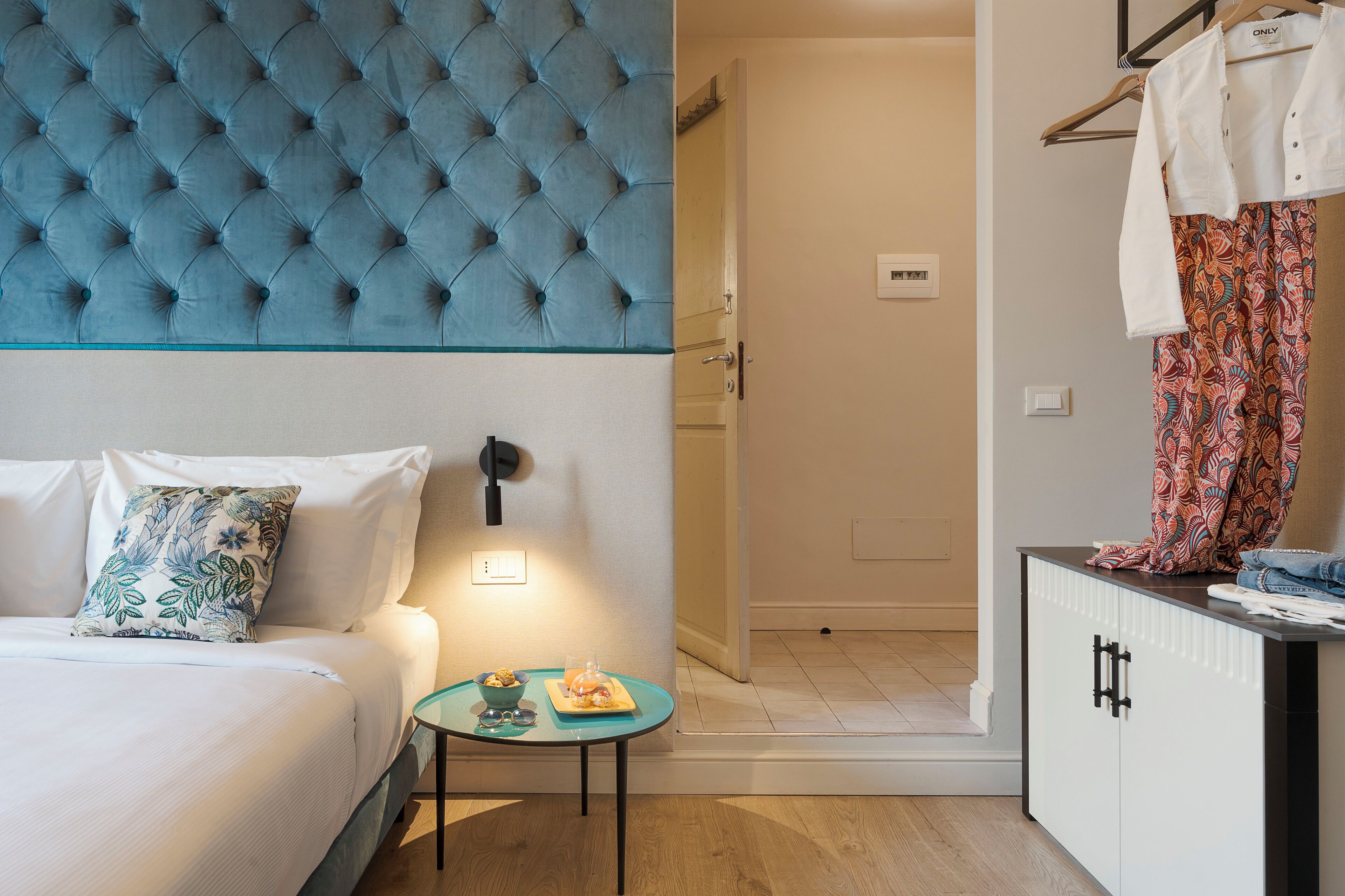 A hotel room with a tufted headboard, a bed with white bedding, a small table with a bowl and a glass, and a wardrobe with patterned curtains.