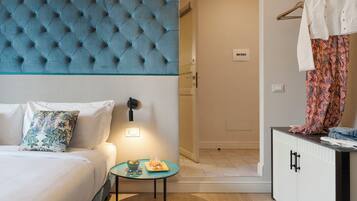 A hotel room with a tufted headboard, a bed with white bedding, a small table with a bowl and a glass, and a wardrobe with patterned curtains.