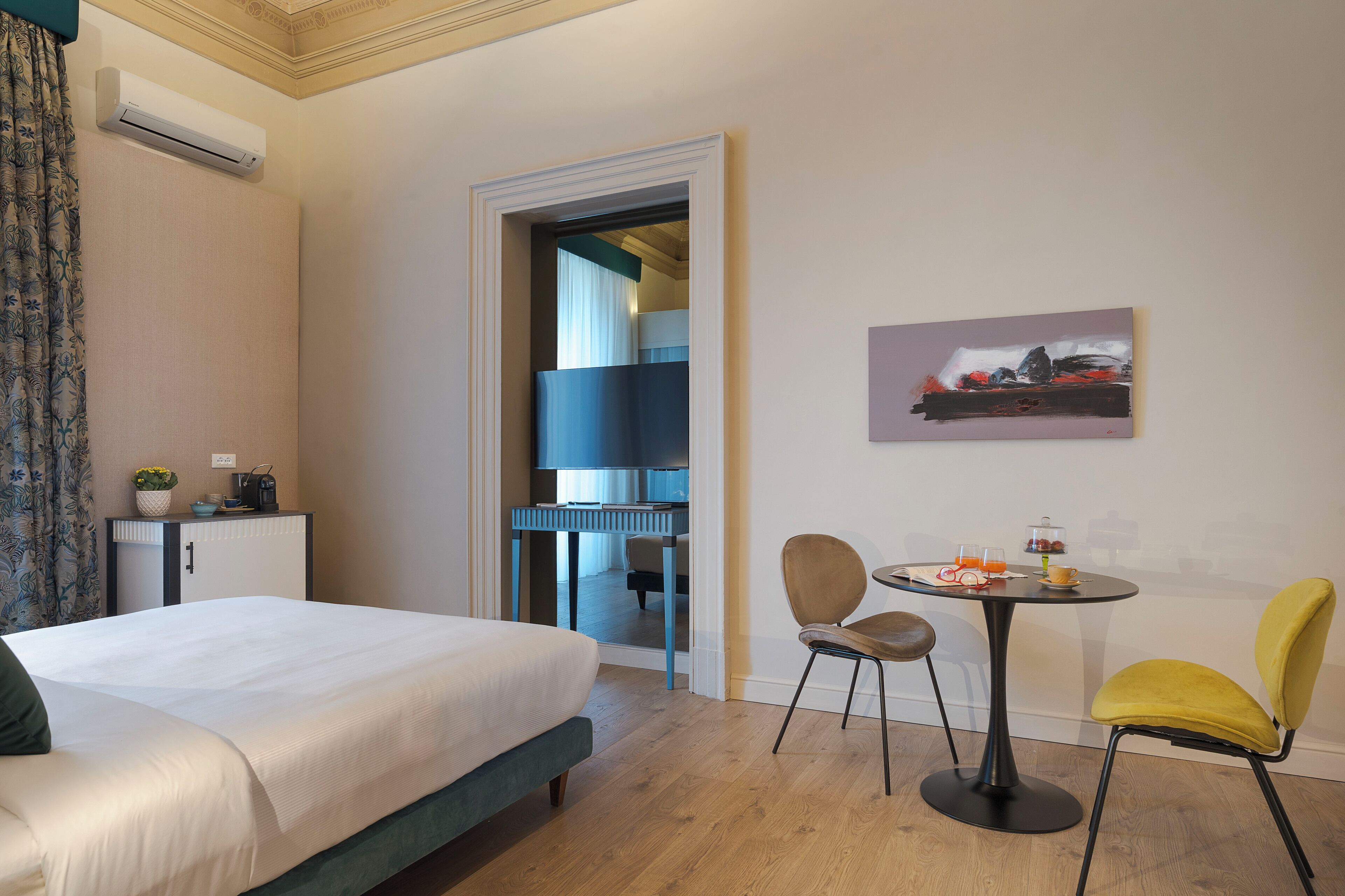 A hotel room with a bed, a desk with a chair, a small table with a chair, and a painting on the wall.