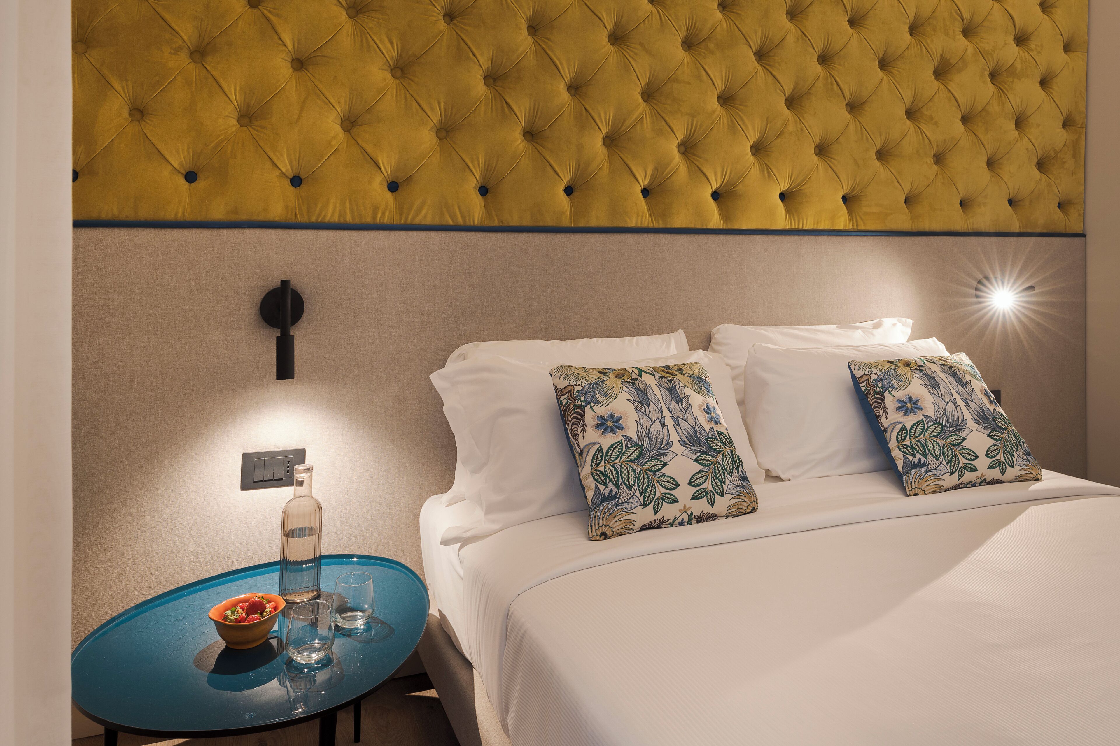 A hotel room with a tufted headboard, a bed with white linens, two floral-patterned pillows, a small table with a bowl of fruit and glasses, and a bottle of water.