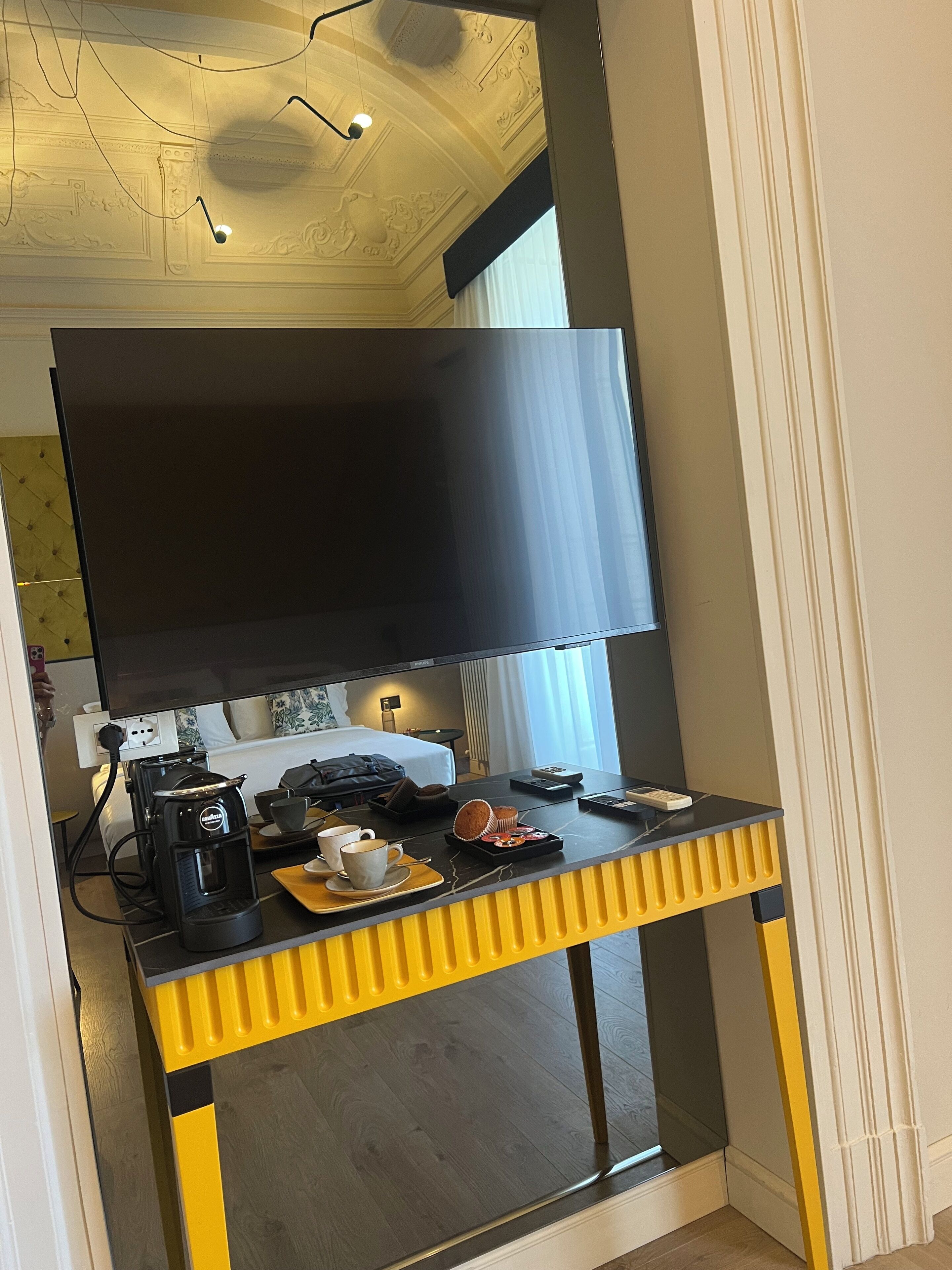 A hotel room with a large flat-screen TV mounted on a wall, a coffee maker, a tray with a muffin, and a remote control on a yellow and black console.