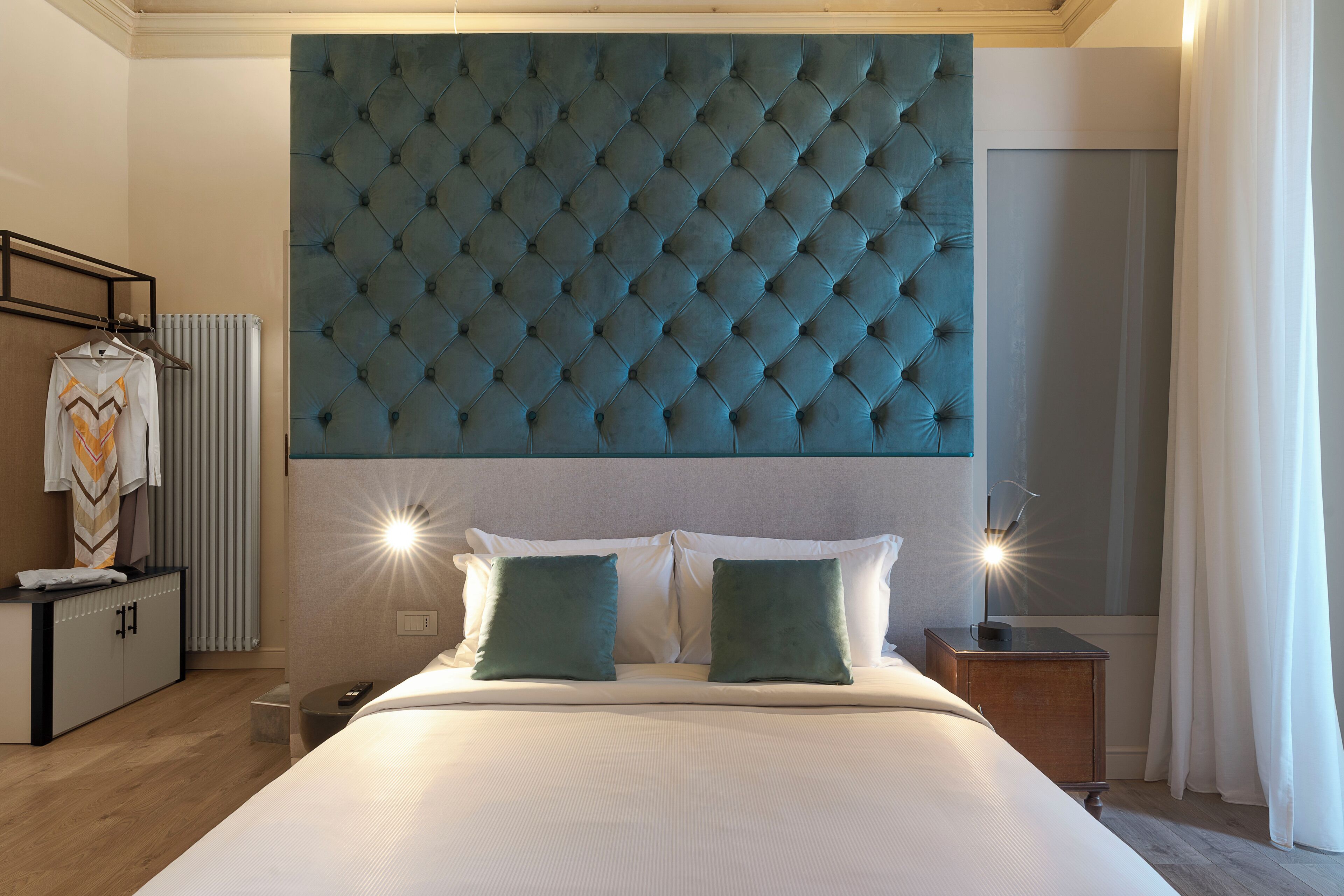 Ein Hotelzimmer mit einem großen, gepolsterten Kopfteil, einem Bett mit weißen Leinentüchern, zwei grünen Kissen, einem Nachttisch mit Lampe und einem Kleiderschrank mit Kleidung.
