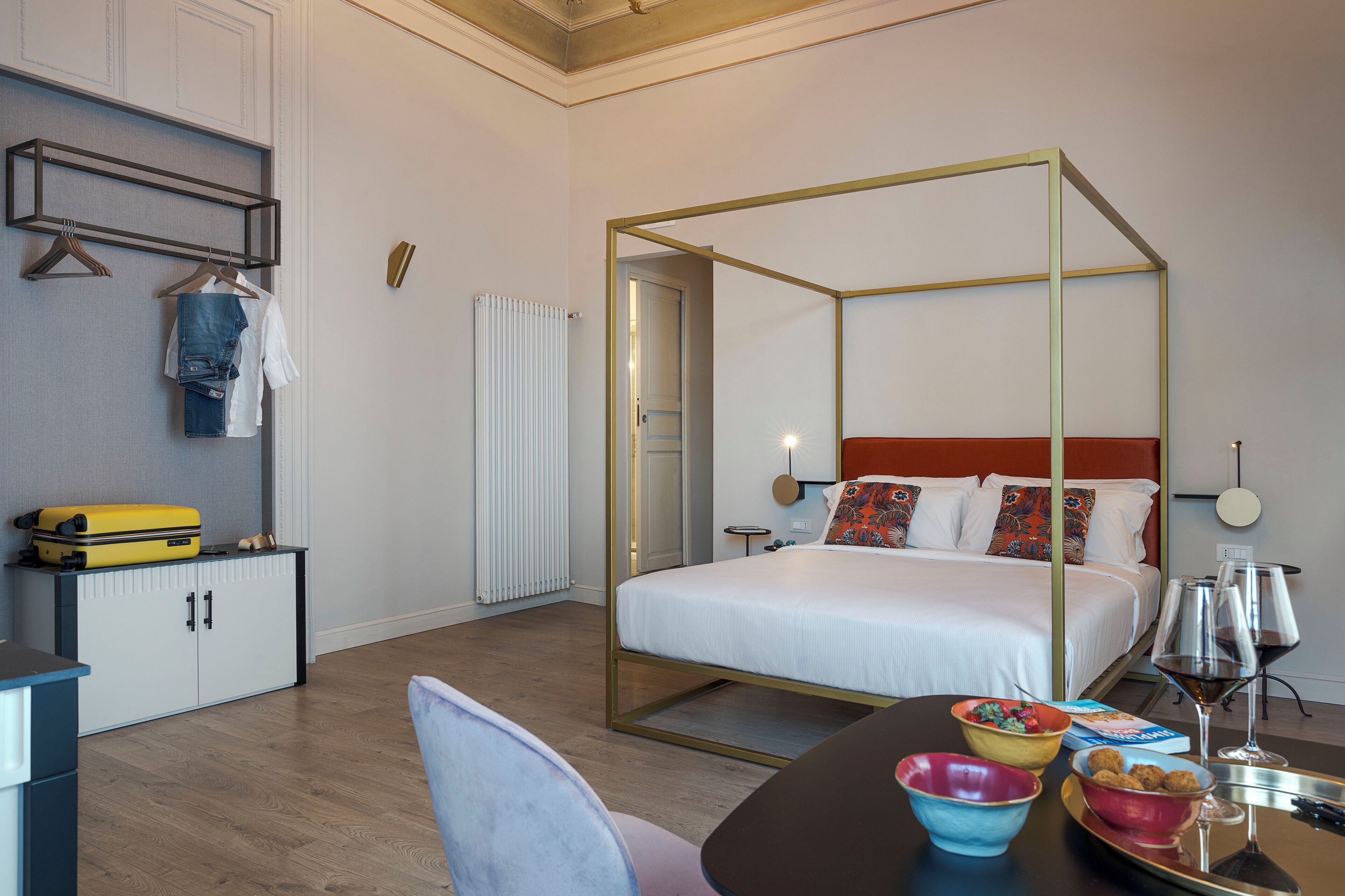 A hotel room with a four-poster bed, a desk with a suitcase, a chair, and a dining table with bowls and wine glasses.