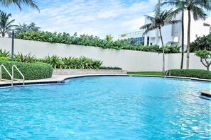 A swimming pool with a curved edge, surrounded by a low white wall and lush greenery, including palm trees and bushes. - Elegant Beachside Resort-Style Condo in Hollywood (Hollywood)
