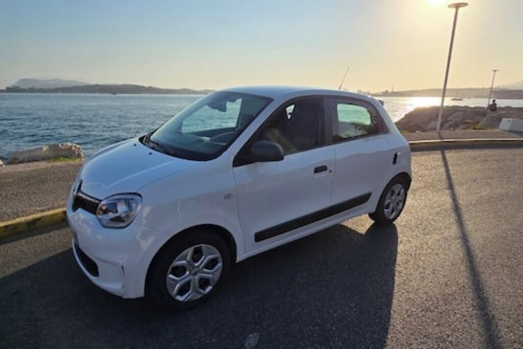 A white car parked by the seaside with a clear view of the ocean and distant hills.