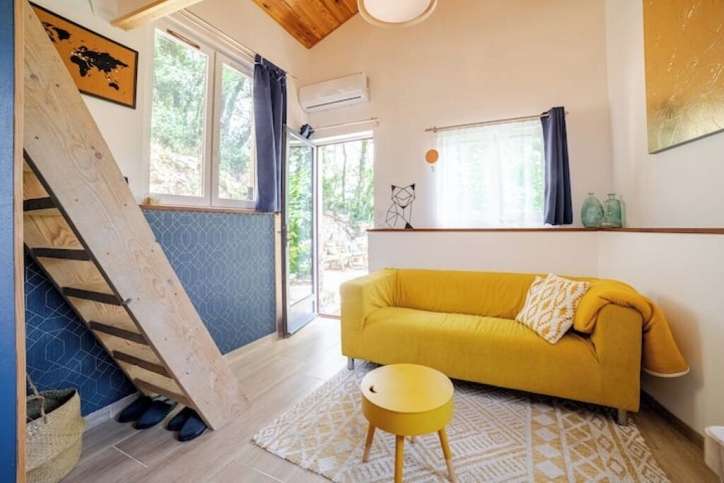 Una sala de estar moderna con un sofá amarillo, una mesita redonda, una escalera y ventanas grandes.