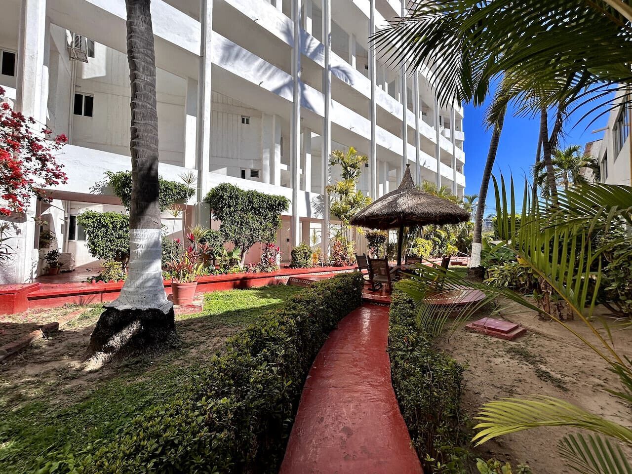 A hotel courtyard with a central pathway, palm trees, and a thatched gazebo.