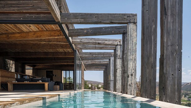 Ein Pool ohne Ende mit Holzbalken darüber, eine Sitzecke mit Kissen und Blick auf die Landschaft.