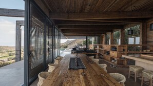 Ein geräumiges Zimmer mit einem großen Holztisch, Korbstühlen und Blick auf eine gebirgige Landschaft durch große Fenster.