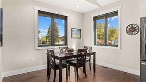 A dining area with a wooden table and chairs, large windows with a view of trees and buildings, and a clock on the wall.