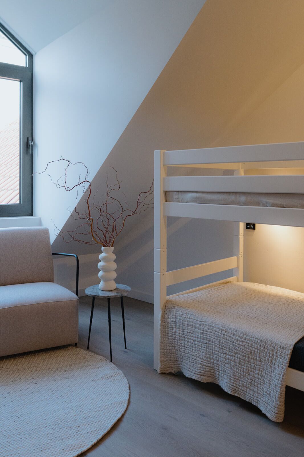A modern bedroom with a bunk bed, a small round table with a vase, a sofa, and a view of the outside through a window.