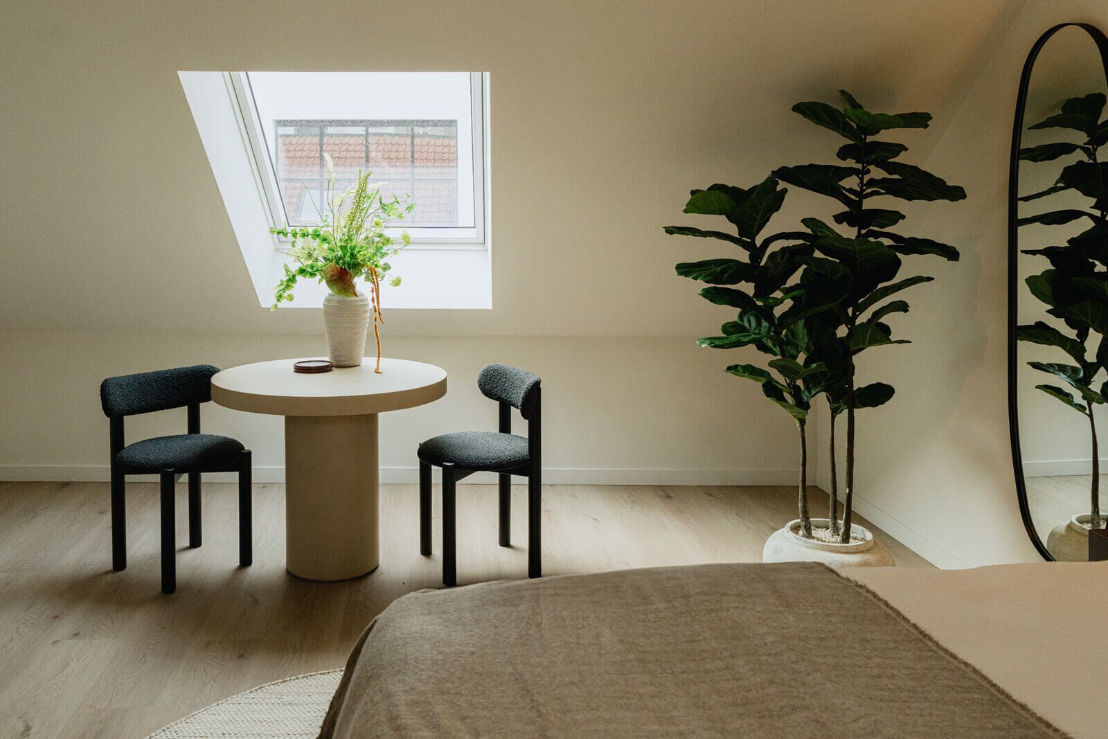 A room with a round table, two chairs, a potted plant, a large mirror, and a bed.
