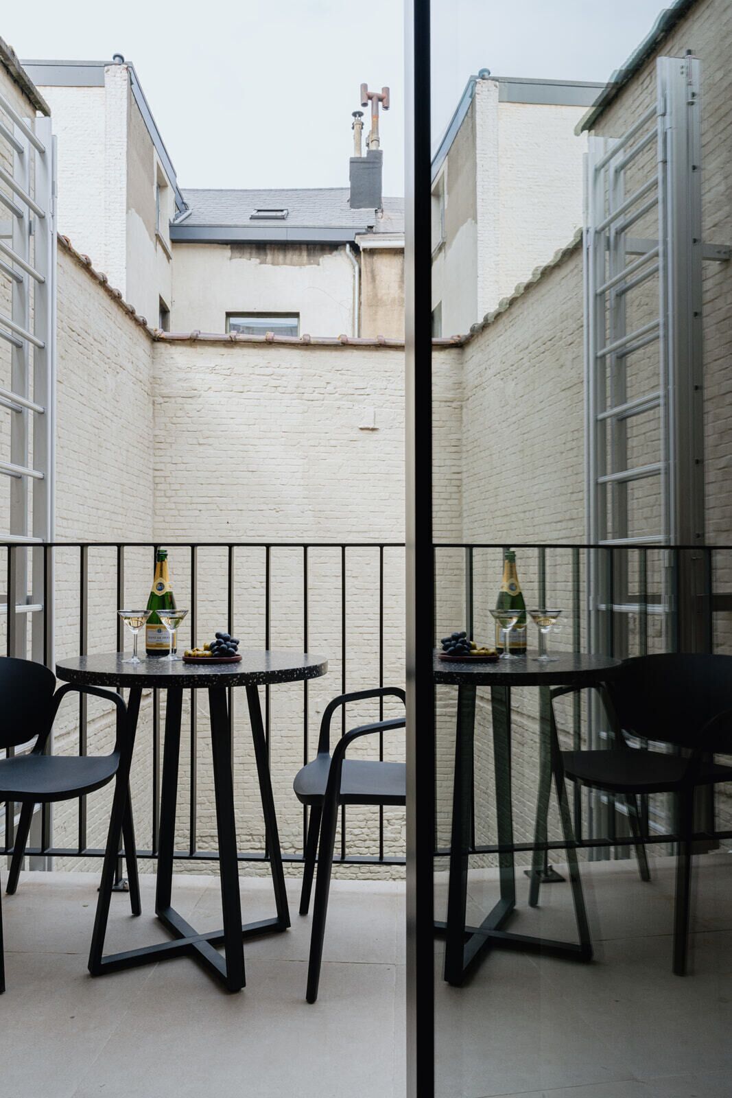 A narrow outdoor patio with two tables and chairs, a bottle of champagne, and a glass on each table.