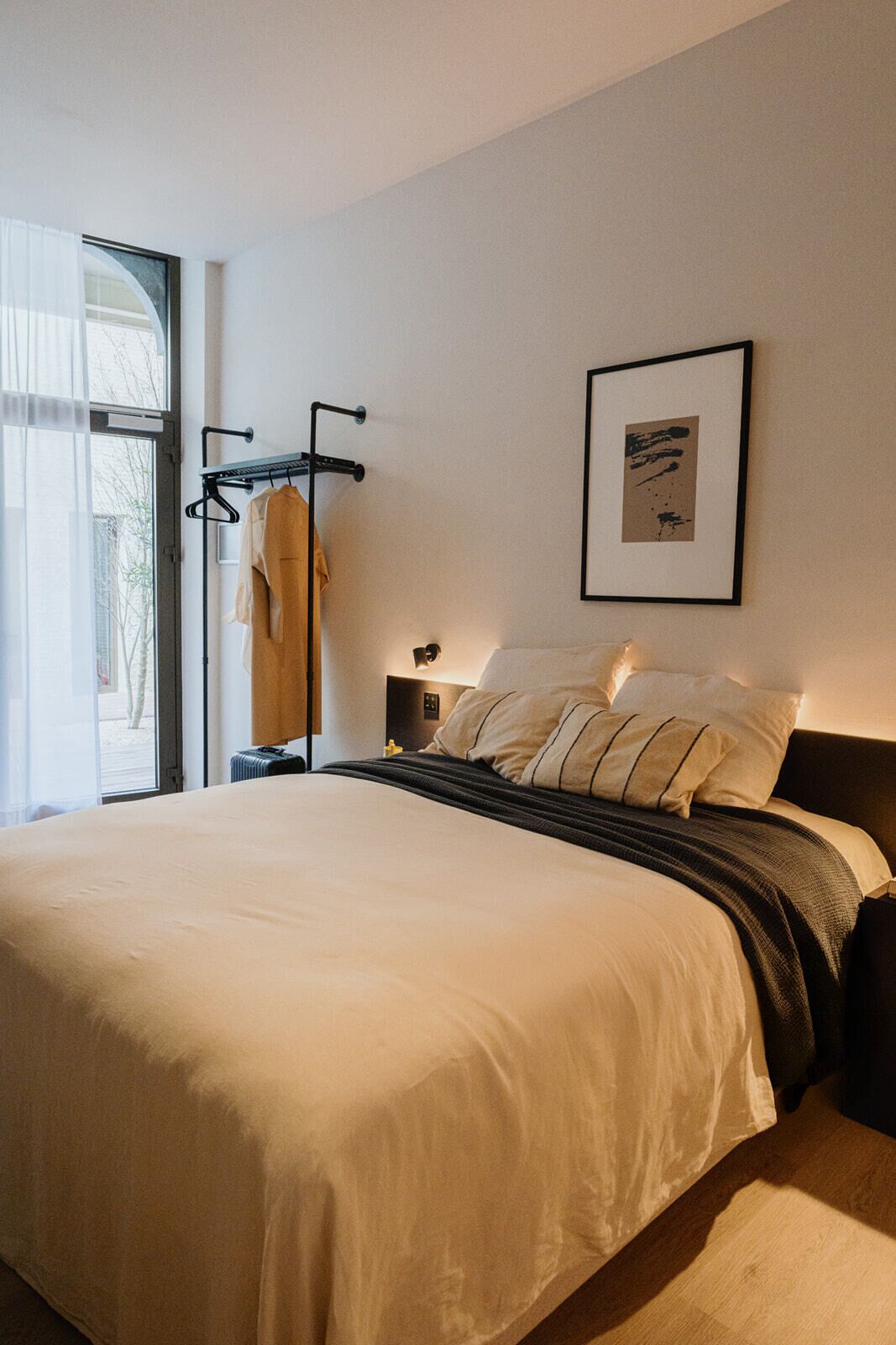 Et moderne hotelværelse med en stor seng, et natbord, et indrammet kunstværk på væggen og et vindue med lette gardiner.
