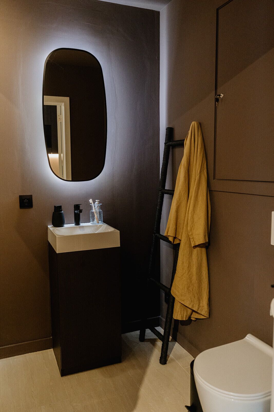 Un baño con una pared oscura, un lavabo blanco, un espejo, un perchero con una toalla amarilla y un inodoro blanco.