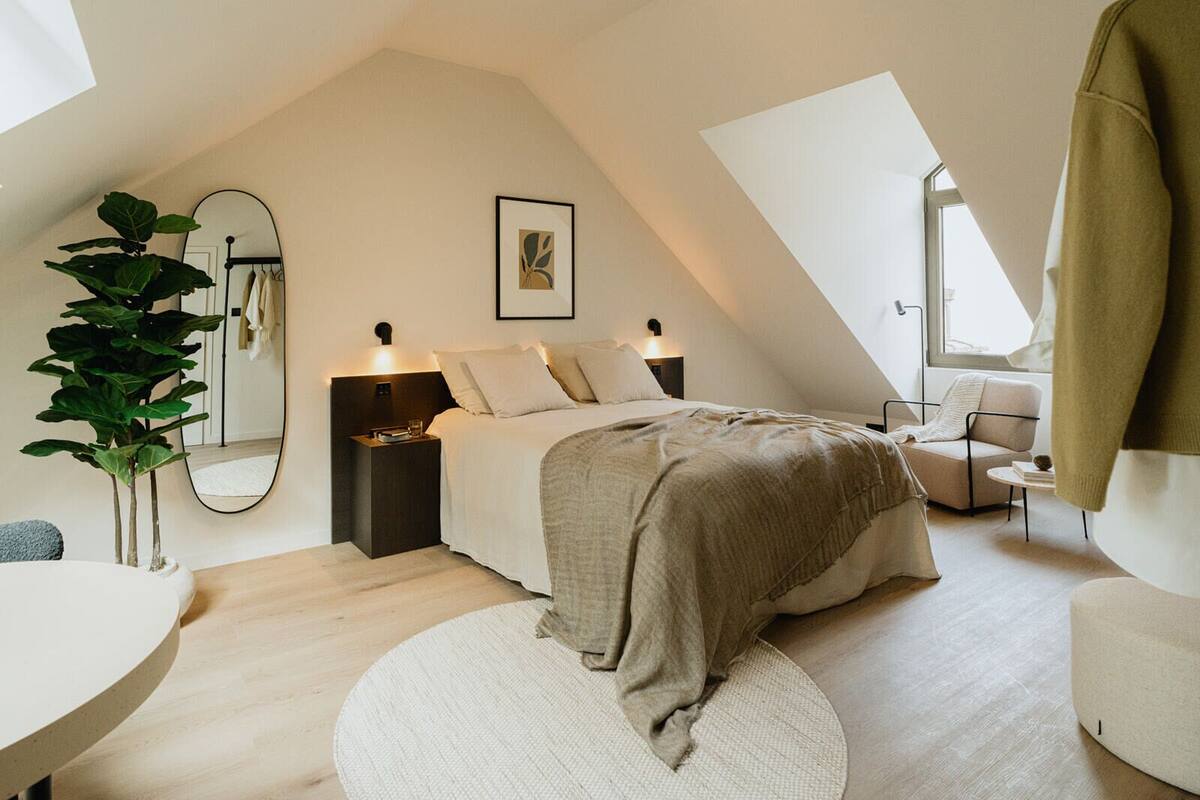 Une chambre moderne avec un plafond en pente, un grand lit, des tables de chevet, un miroir, une plante et un coin salon avec une chaise et une table.
