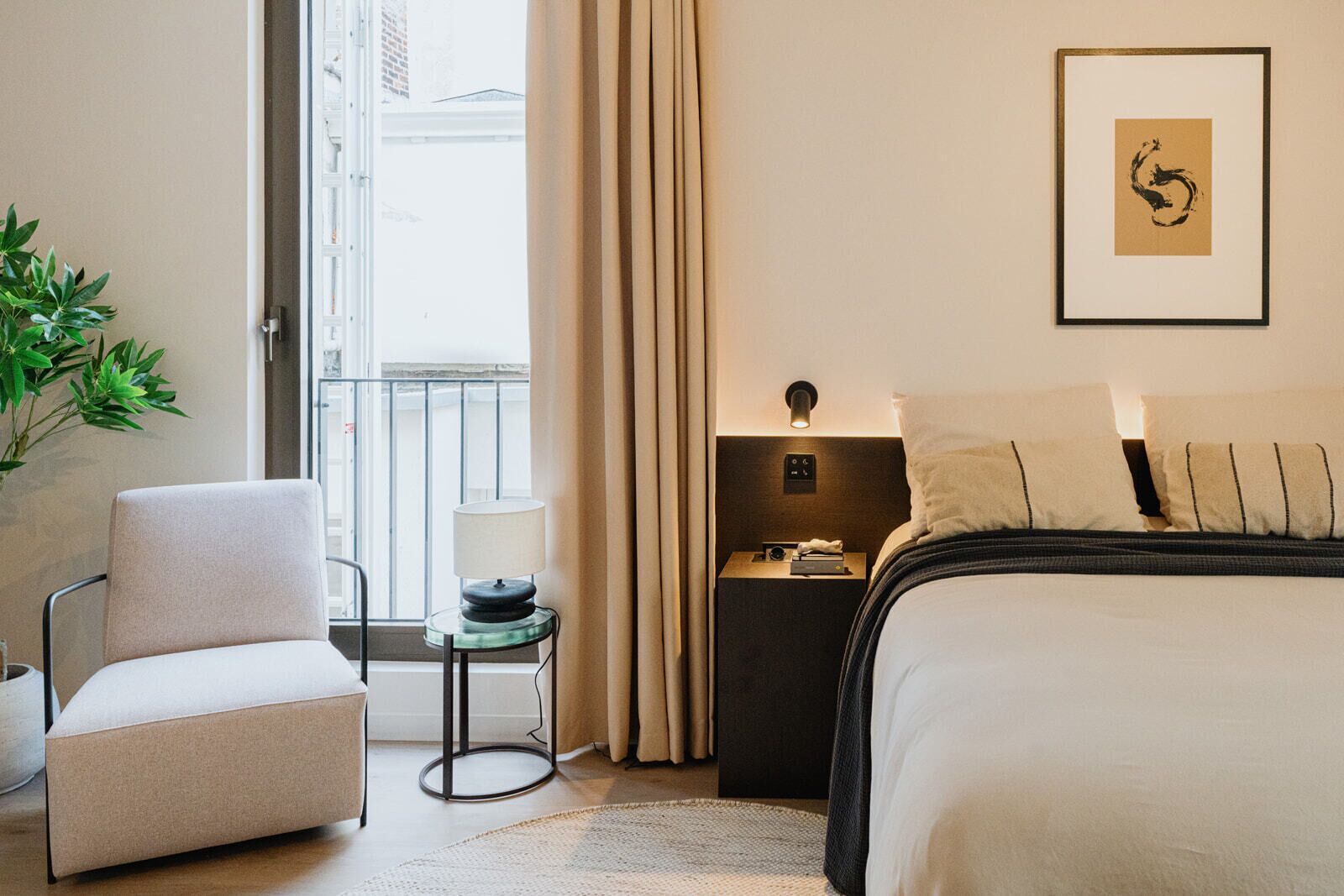 A hotel room with a bed, a chair, a nightstand, a lamp, and a framed artwork on the wall.
