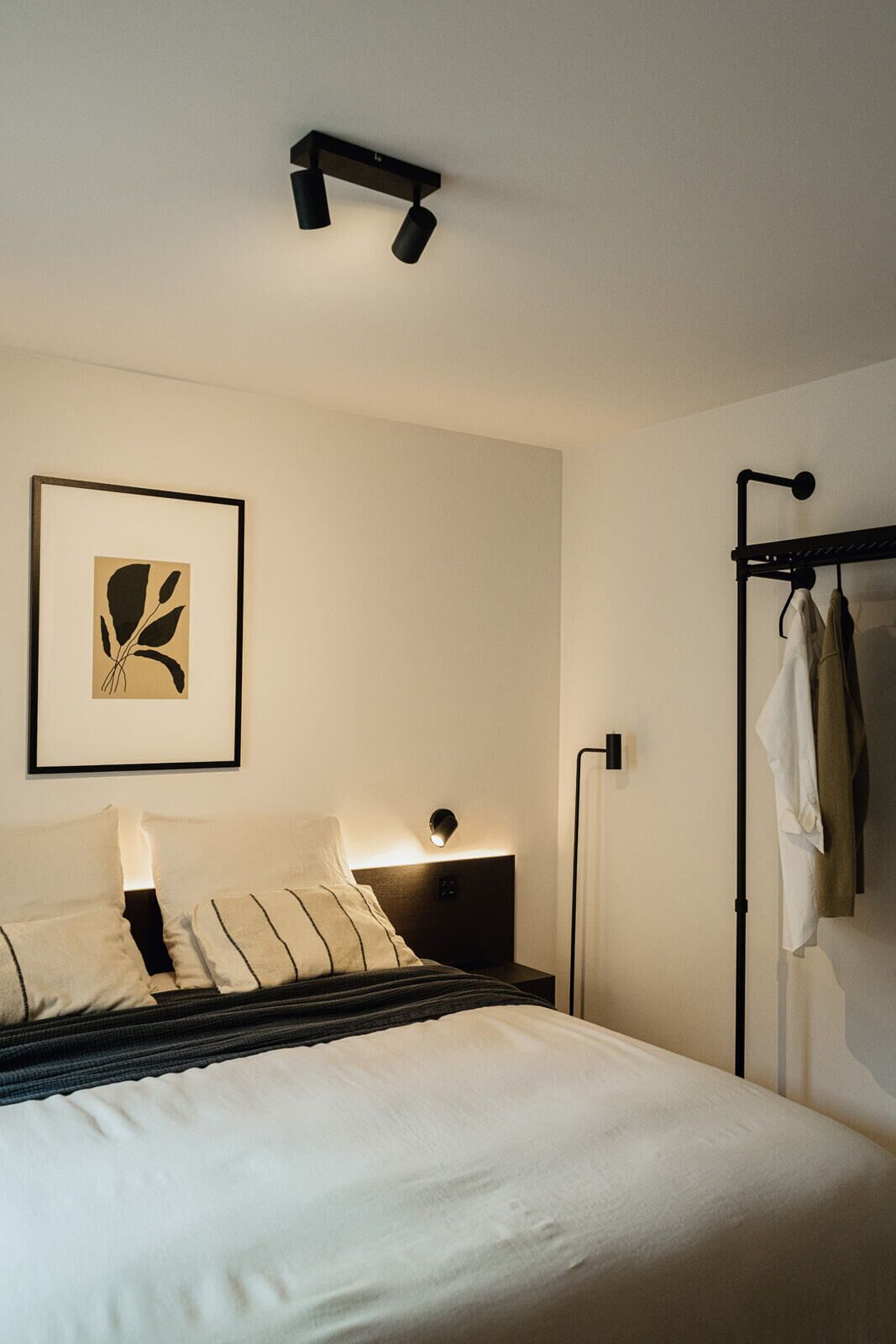 Ein ordentlich bezogenes Bett mit Kopfteil, einer Nachttischlampe und einem gerahmten Kunstwerk an der Wand.