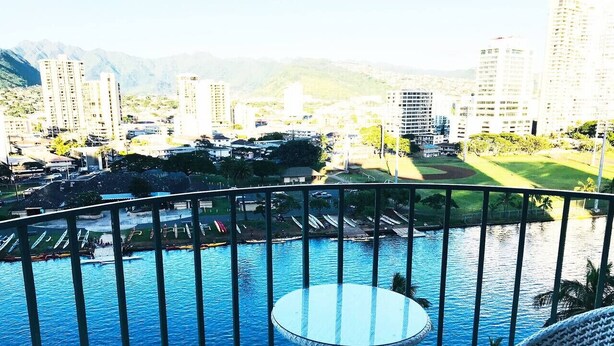 A balcony with a view of a swimming pool, surrounded by high-rise buildings and greenery. - Waikiki 2 bedrm Condo Canal View w/ Gated Parking (Honolulu)
