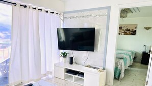 A modern hotel room with a large flat-screen TV mounted on a marble wall, a white console table with a remote control, a small potted plant, and a view of the city through the window. - Waikiki 2 bedrm Condo Canal View w/ Gated Parking (Honolulu)