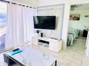 Ein modernes Hotelzimmer mit einem großen Flachbildfernseher an einer Marmorwand, einem weißen Konsolentisch mit Fernbedienung, einer kleinen Topfpflanze und Blick auf die Stadt durch das Fenster.