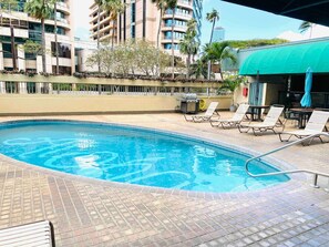 Ein Swimmingpool mit Liegestühlen und Sonnenschirmen, umgeben von einer Ziegelterrasse und Palmen.