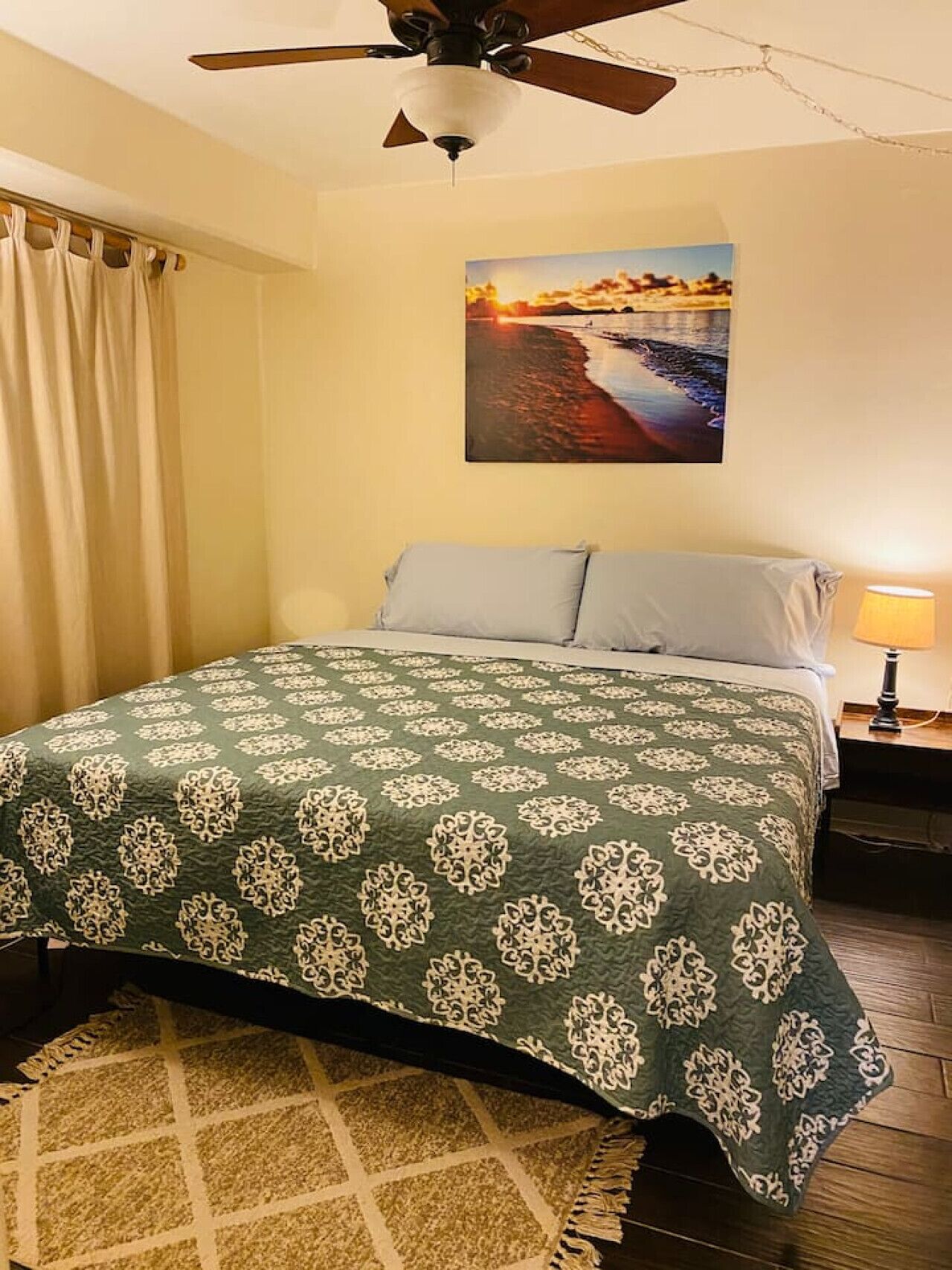 A bedroom with a bed, a ceiling fan, a painting of a sunset, and a nightstand with a lamp.