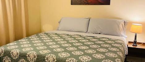 A bedroom with a bed, a ceiling fan, a painting of a sunset, and a nightstand with a lamp.