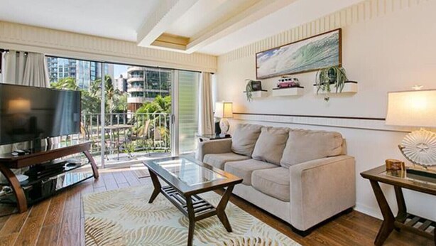 A modern living room with a sofa, coffee table, TV, and balcony with a view of buildings and greenery. - Waikiki 2 Bedroom w/ Covered Parking, Family Condo (Honolulu)