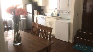 Una cocina pequeña con armarios blancos, una estufa y un lavamanos. Hay una silla de madera y un florero con flores sobre la mesa.