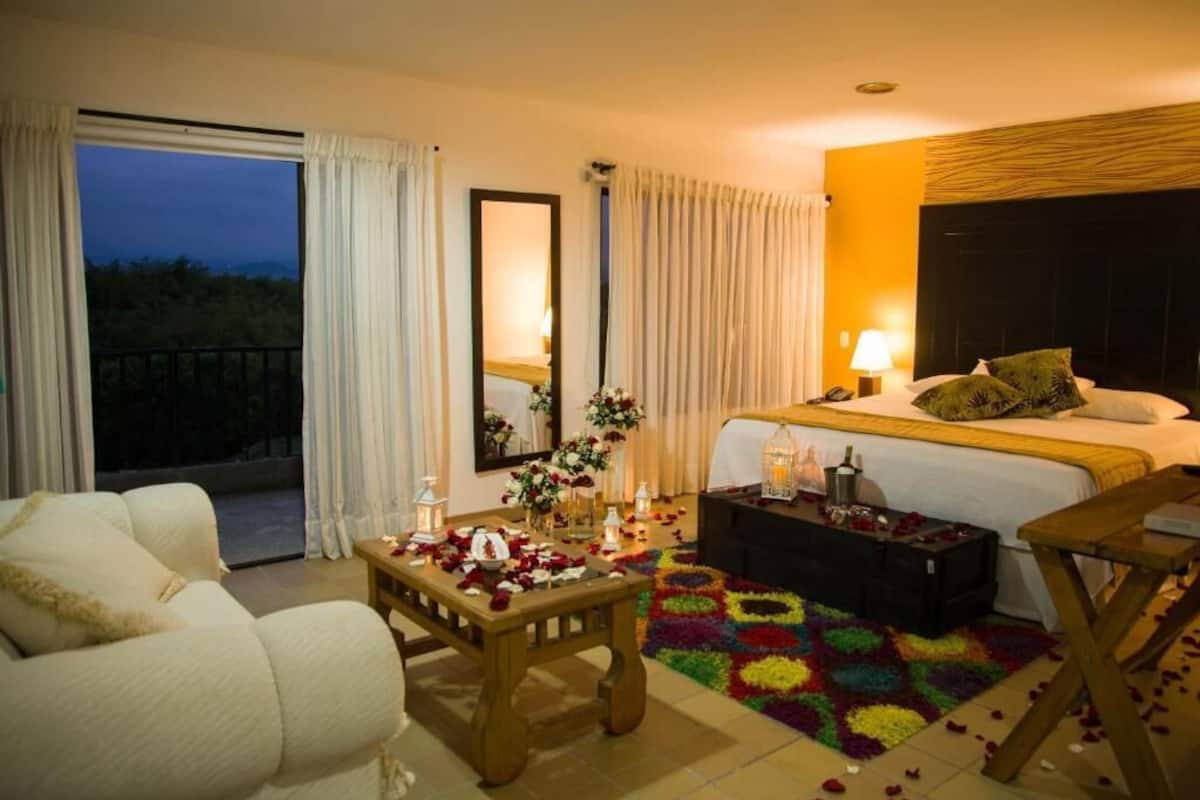 Una camera d'albergo con un letto, una poltrona, un tavolino con petali di rosa, un tappeto colorato e un balcone con tende.
