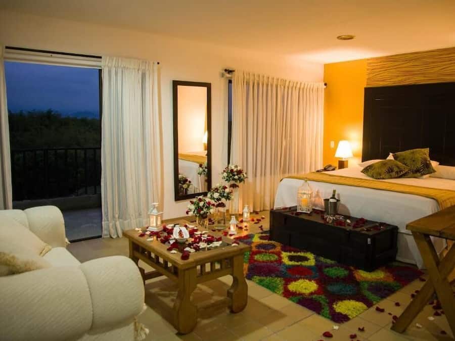 Quarto de hotel com cama, sofá, mesinha com pétalas de rosa, tapete colorido e varanda com cortinas.