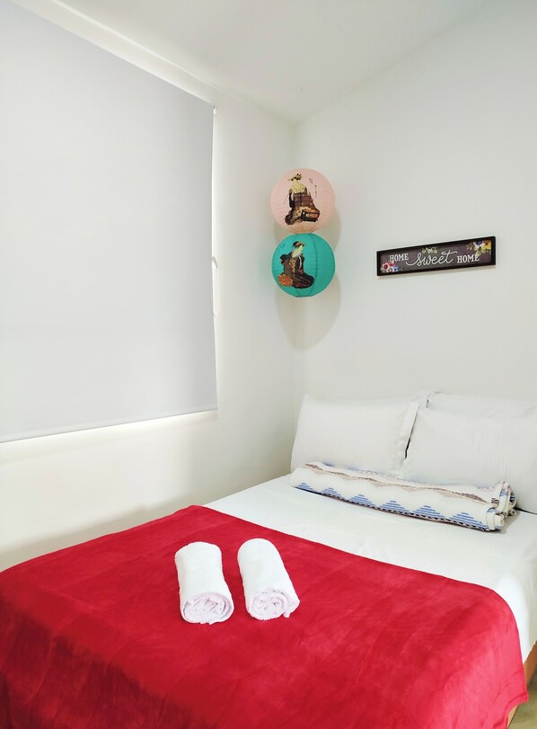 Cama bem arrumada com um cobertor vermelho e duas toalhas enroladas, uma parede branca e duas decorações circulares na parede.