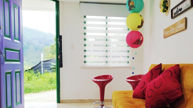 Um quarto com uma porta roxa, um sofá amarelo com almofadas vermelhas e uma janela com persianas.