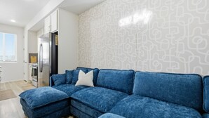 Een moderne woonkamer met een blauwe modulaire bank, een keukenhoek met een koelkast, en een behang met een patroon.